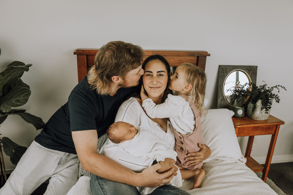 dad and girl kissing mum on cheeks while she is looking at the camera holding her baby