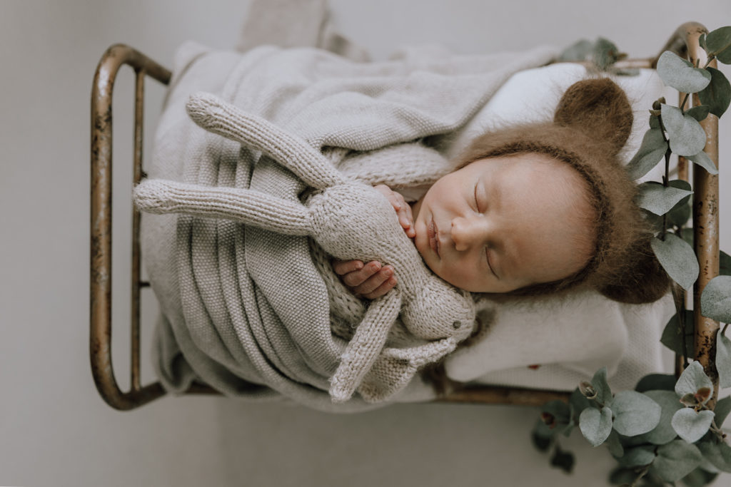 baby boy with a brown teddy hat holding a beige bunny in a brass bed with a neutral background