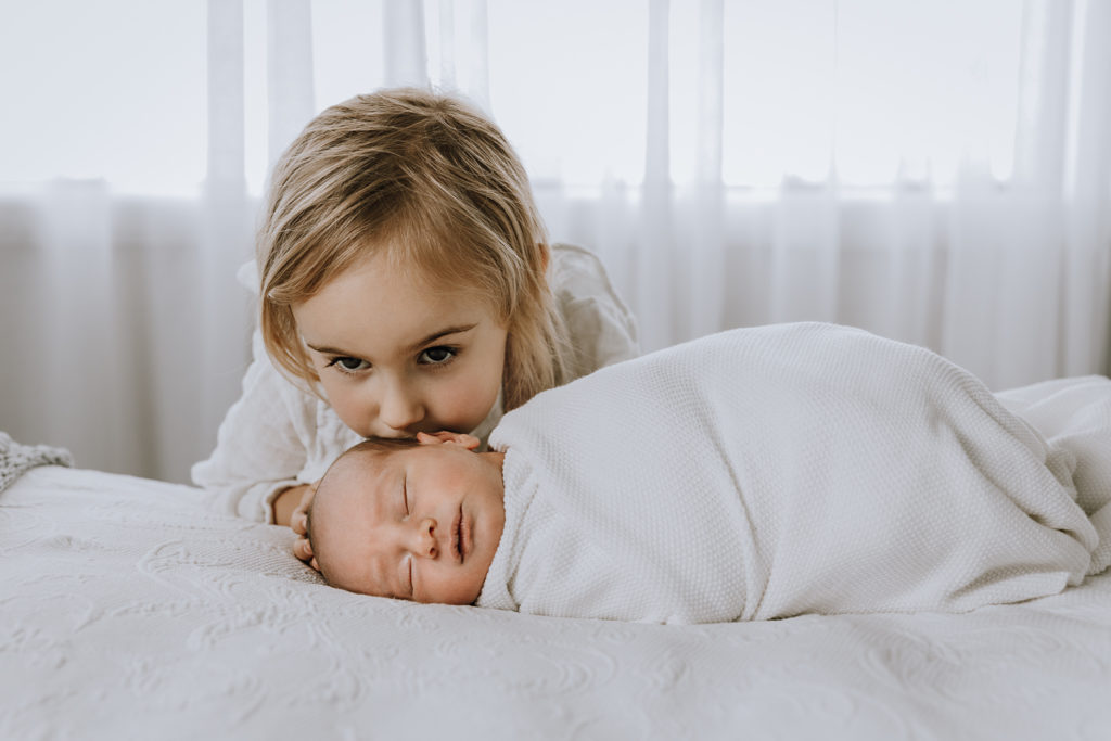 siter kissing baby brothers head on a white bed in front of a window