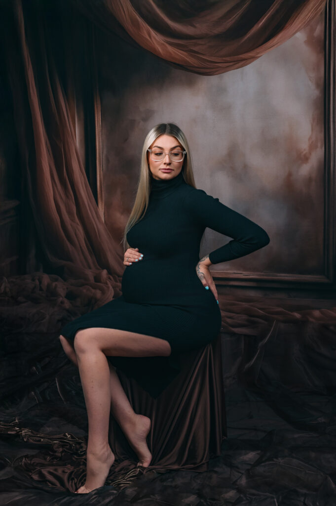 blonde pregnant lady in simple black dress seated infront of an elaborate draping fabric and frame backdrop