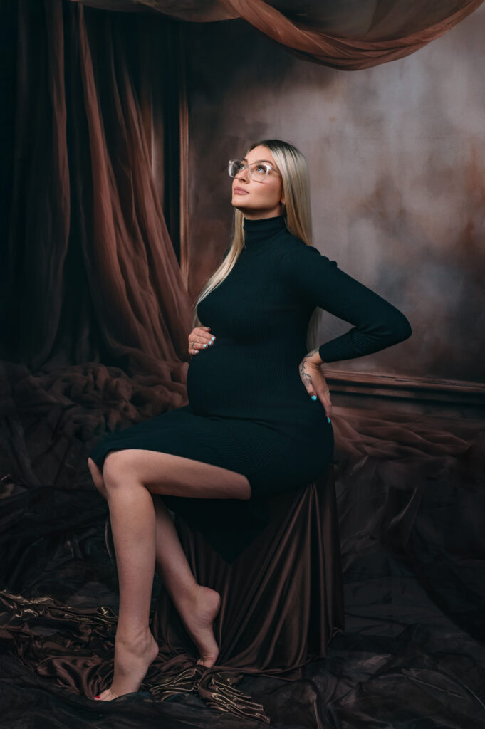 blonde pregnant lady in simple black dress seated infront of an elaborate draping fabric and frame backdrop