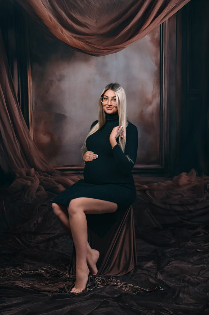blonde pregnant lady in simple black dress seated infront of an elaborate draping fabric and frame backdrop, smiling t camera
