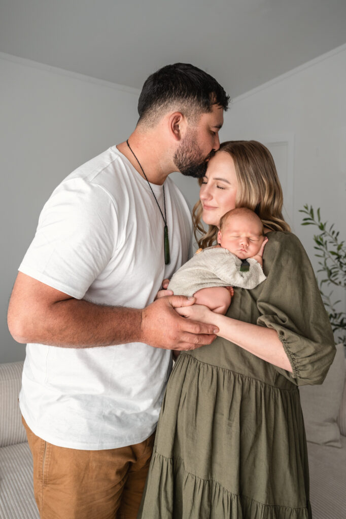 mum holding sleeping baby on shoulder with dad hugging in and kissing her on top of the head