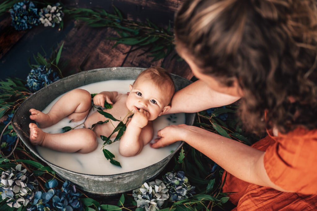 baby boy and his Mum holding him in a tin bath with blue hydrangea flowers and gum leaves enjoying his milk bath getting photographed


